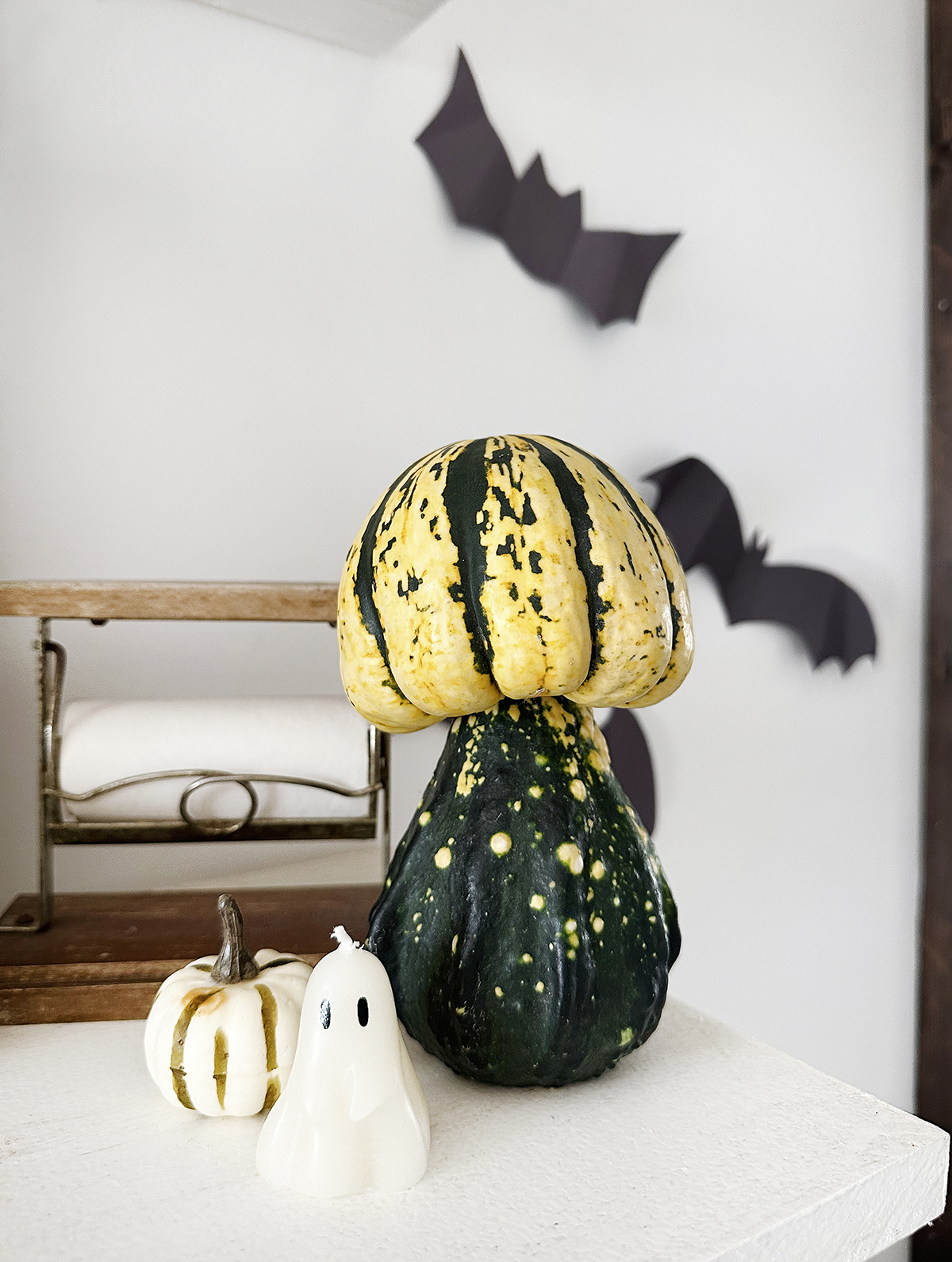 stacked pumpkins to look like mushrooms on white shelf next to ghost candle with cut out bats on wall in background